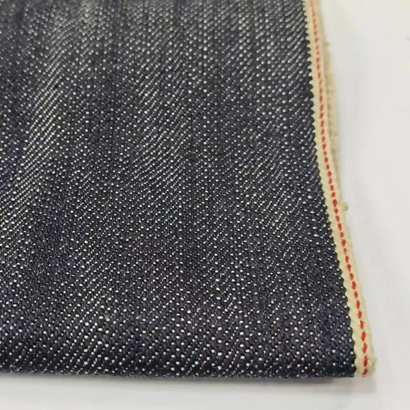 Un gros plan du tissu en jean selvedge 16,5 oz de Longan Craft France (100 % coton) révèle un sergé bleu foncé, des fils de trame blancs et un bord fini avec une bande crème et une ligne rouge. Disponible au mètre comme "Tissu en jean selvedge 16,5 oz.