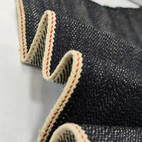 Un gros plan du tissu en jean selvedge 16,5 oz au mètre (100 % coton) de Longan Craft France révèle sa surface texturée, sa lisière crème et ses coutures rouges, avec le denim lourd plié doucement sur un fond blanc.