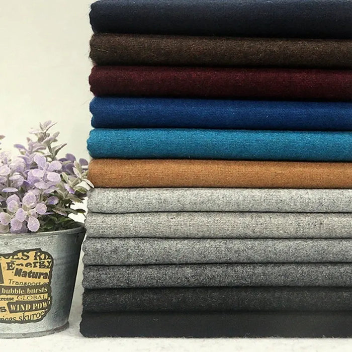 Une pile de tissu flanelle en laine au mètre de Longon Craft France, disponible dans les tons noir, brun, bleu, tan et gris, est présentée à côté d'un pot avec des fleurs violettes claires - idéal pour les vêtements d'hiver ou le textile d'intérieur cosy.